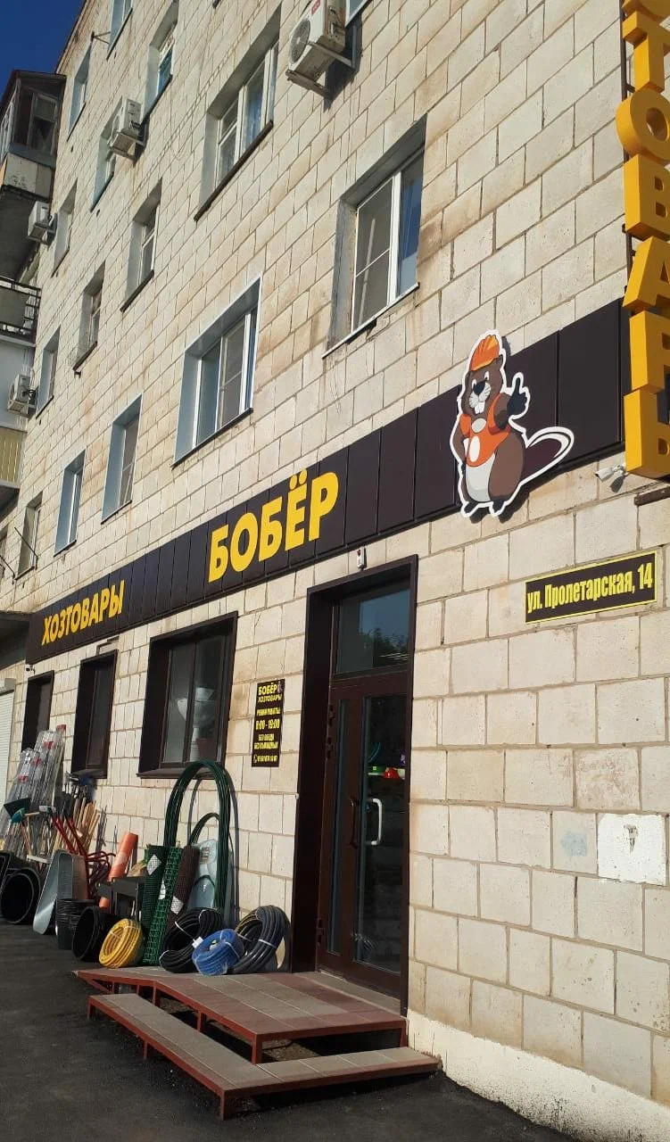 Вход в магазин «Хозтовары Бобёр»: коричневая вывеска с жёлтыми буквами, у двери — шланги и садовый инвентарь